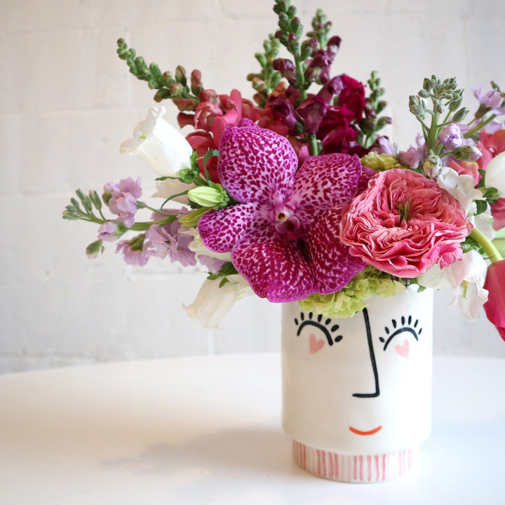 Face vase, with heart eyes a nose and a smile. Filled in the vase is burgundy snapdragons, pink tulips, light purple stock, white campanula and fushia orchid on a white background.