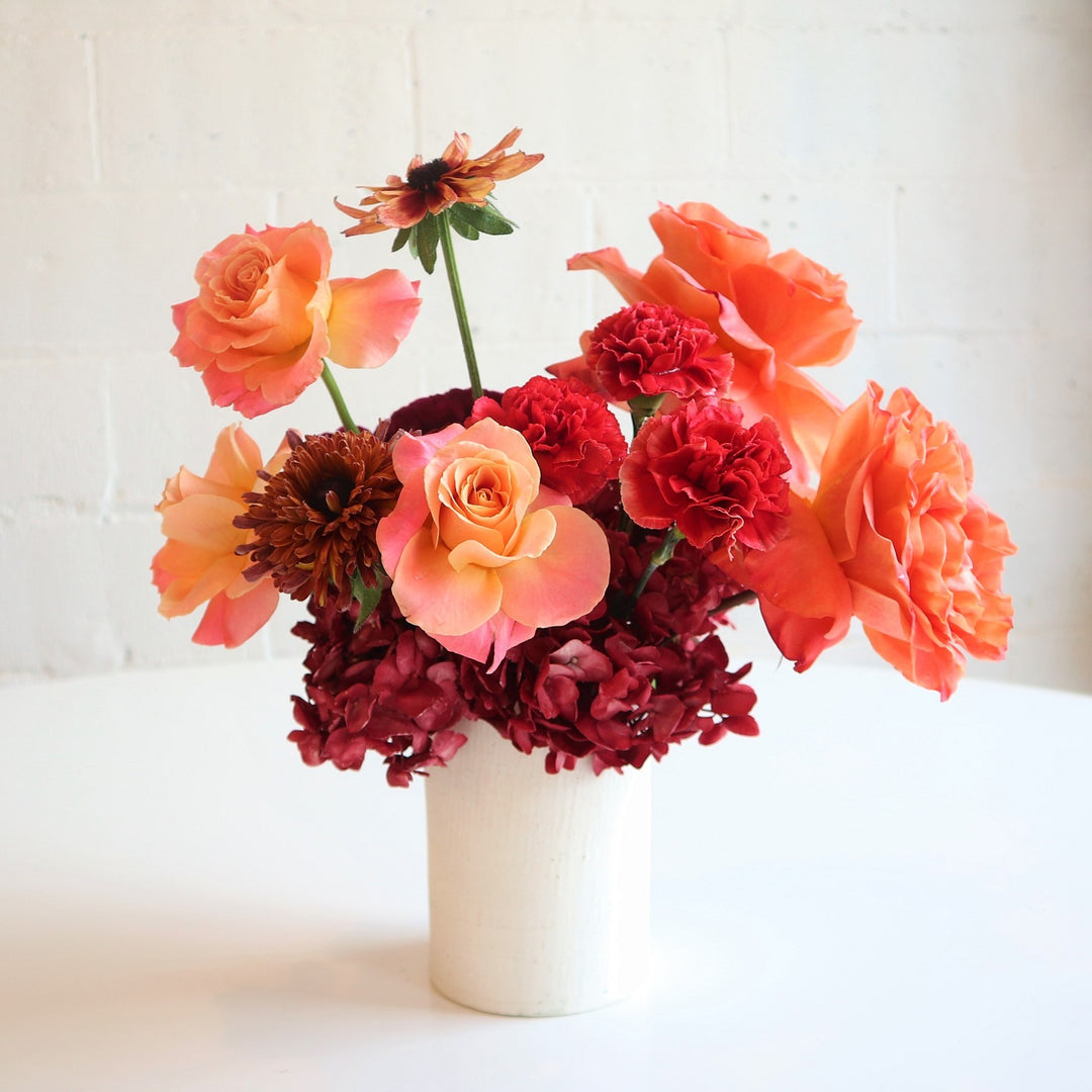 Abundance | A perfect mix of summer and fall. This arrangement contains reds, oranges, and peach. In a white vase, focusing on roses, mums, and hydrangea.