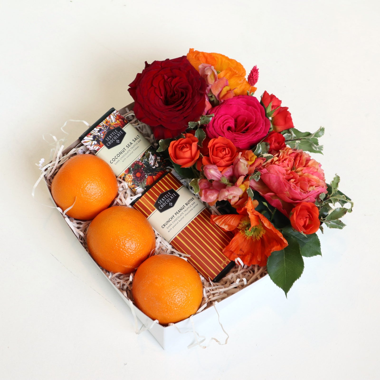 Gift box with two Seattle chocolate bars, three oranges, a floral arrangement with red roses, fushia roses, orange spray roses, orange poppies, varigated pitt on a white background. 