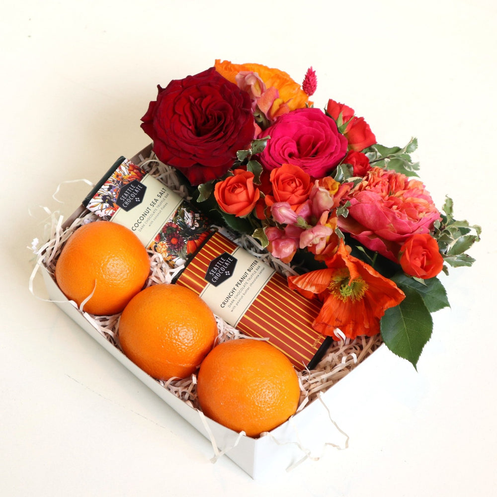 Gift box with two Seattle chocolate bars, three oranges, a floral arrangement with red roses, fushia roses, orange spray roses, orange poppies, varigated pitt on a white background.
