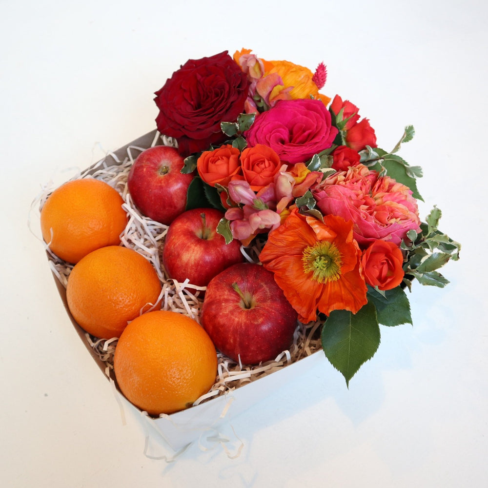 Gift box with a red rose, pink rose, orange spray rose, orange poppies and greenery alongside three apples and three oranges.