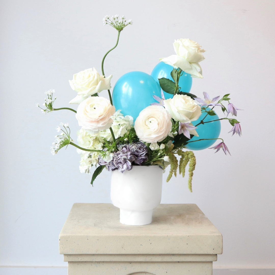 Elegant floral arrangement in a white vase featuring white roses, ranunculus, hydrangeas, and lavender accents, paired with bright blue balloons for a cheerful touch.