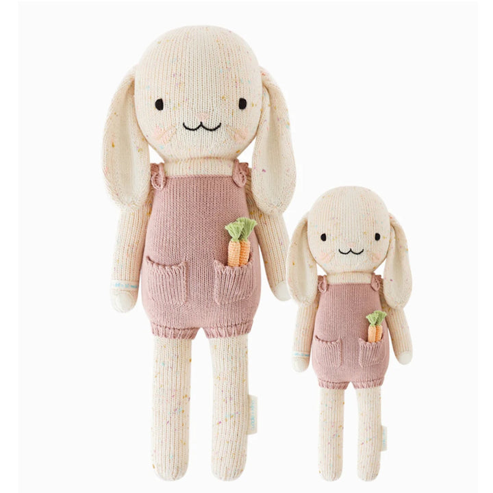 Harper The Bunny | Cuddle + Kind | A subtle confetti pattern on a cream knitted bunny plush that is wearing pink overalls/romper that has two knit carrots in the pocket. Two sizes pictured: 20" and 13".
