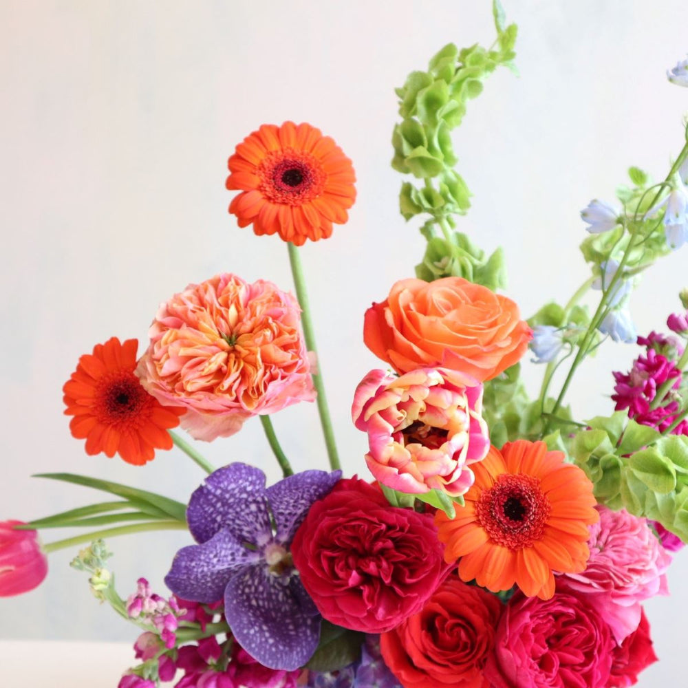 Bright and colorful floral arranegement, with orange gerbera daisies, bells of ireland, fushia garden roses, blue delphinium, purple vanda orchids, burgundy stock in a purple low compote.