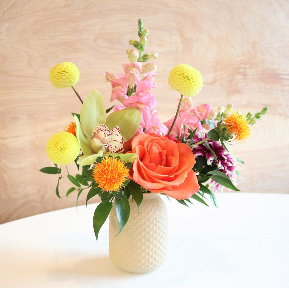Rising Sun | Vased Arrangement | A light colored vase with yellow, green, pink, and orange florals. Accented with greenery. Photo taken with a wooden background.