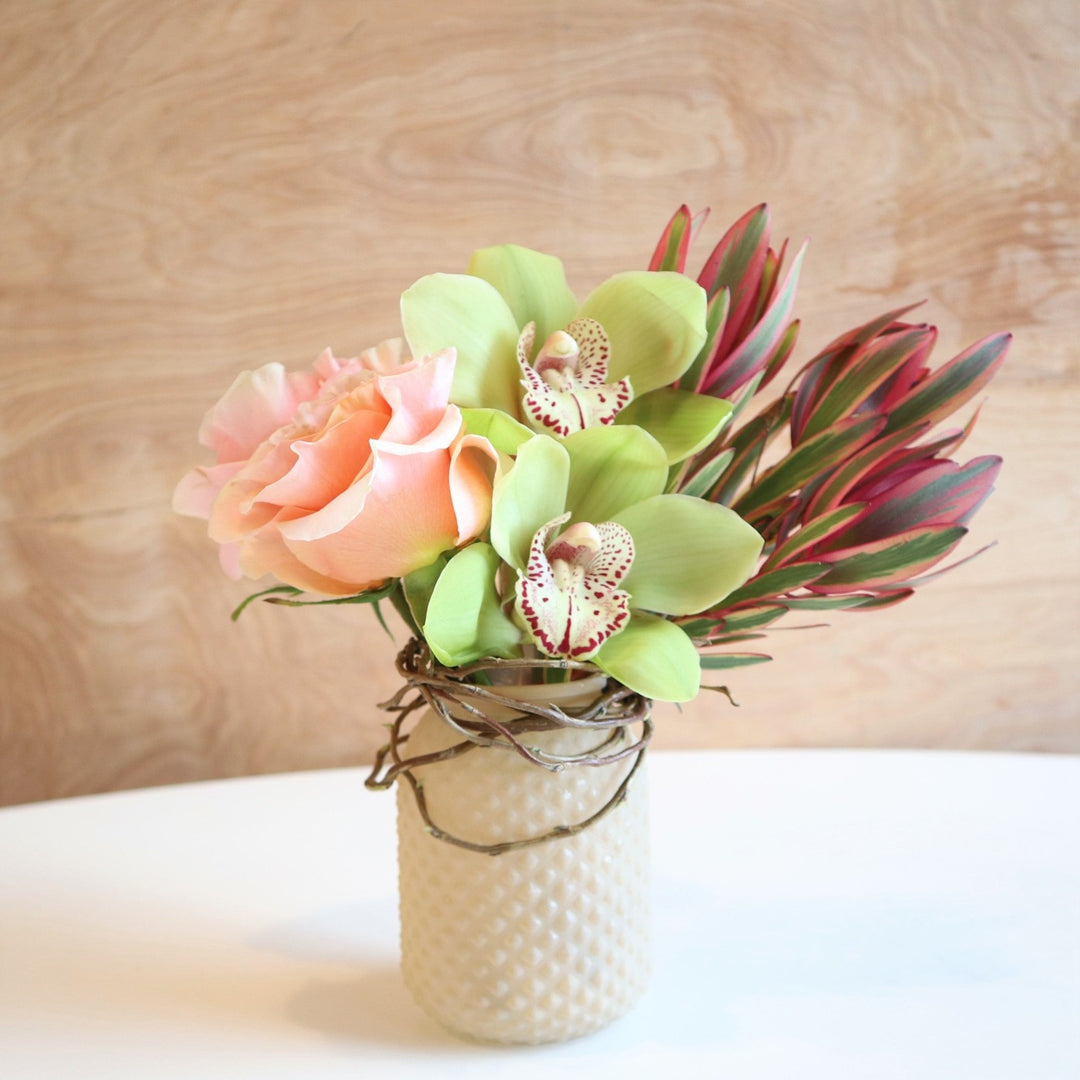 Glimmer | A vased arrangement with roses, orchids, and Leucadendron.  Accent branching is wrapped around the top of the vase. Colors are light pink and green.
