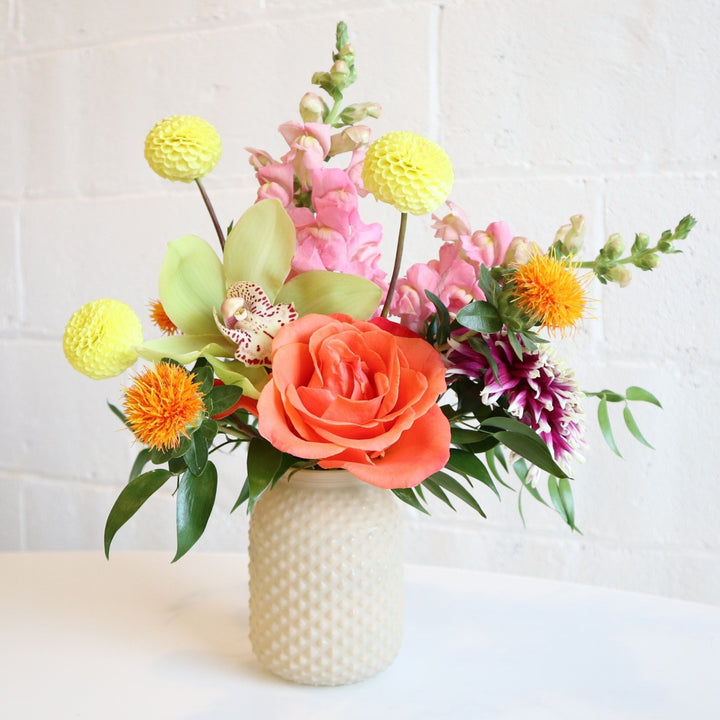 Rising Sun | Vased Arrangement | A light yellow vase with yellow, pink, orange, and accent greenery. Some flowers shown are dahlias, snapdragons, an orchid, and a rose.