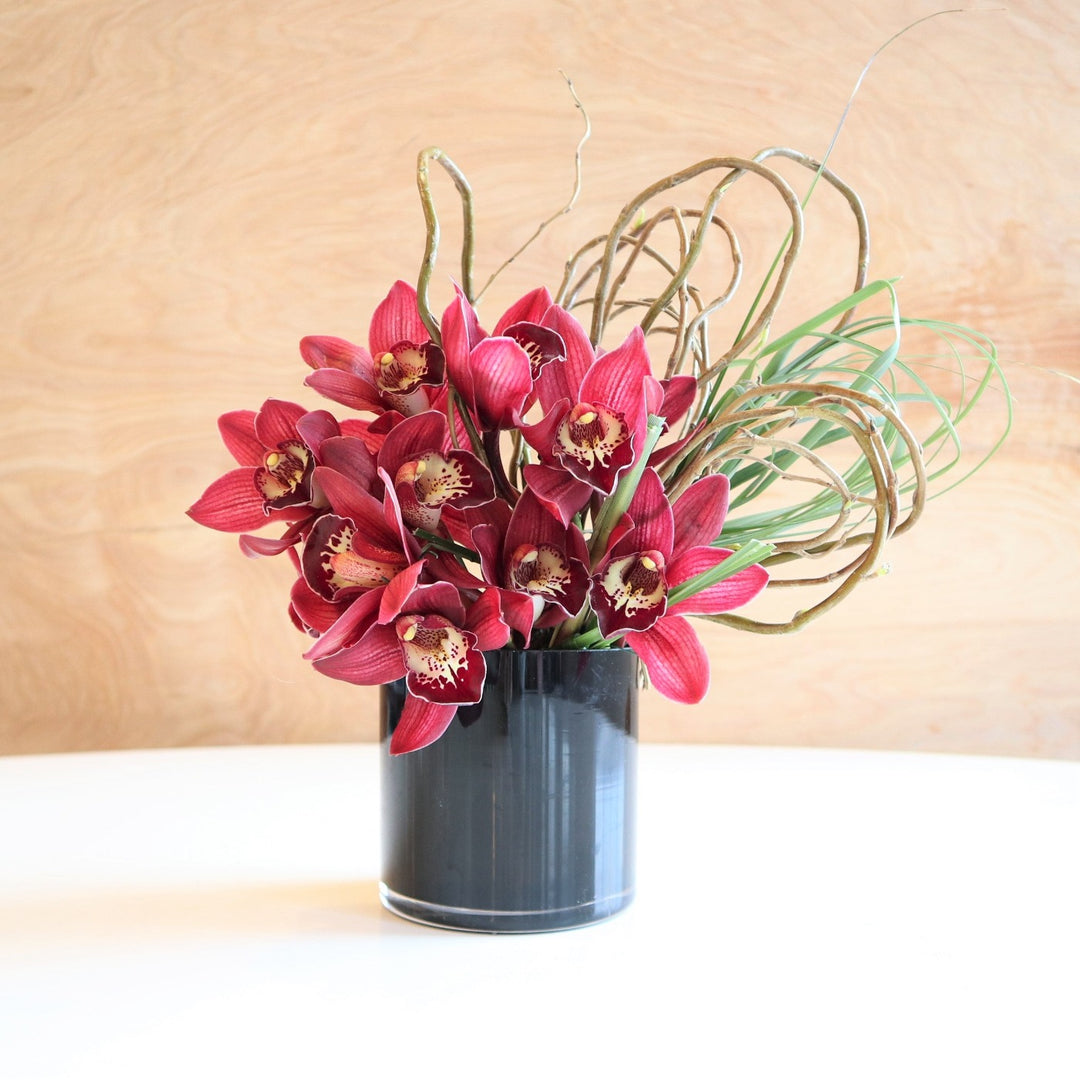 Orchid Delight | Vased Arrangement | A black glass vase with red orchids, branching and grasses.