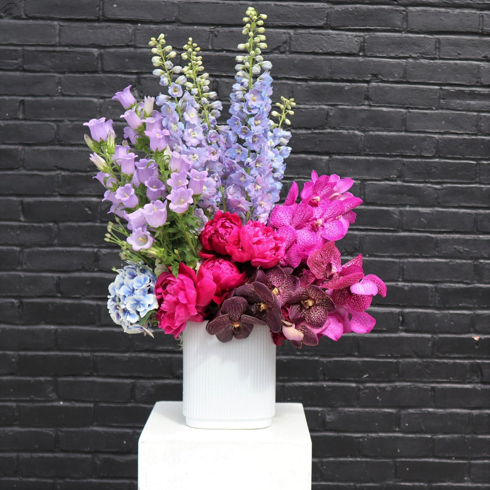 Flower arrangement with magenta and purple orchids, blue delphinium, purple campanula, blue hydrangea and pink peonies in a white vase.