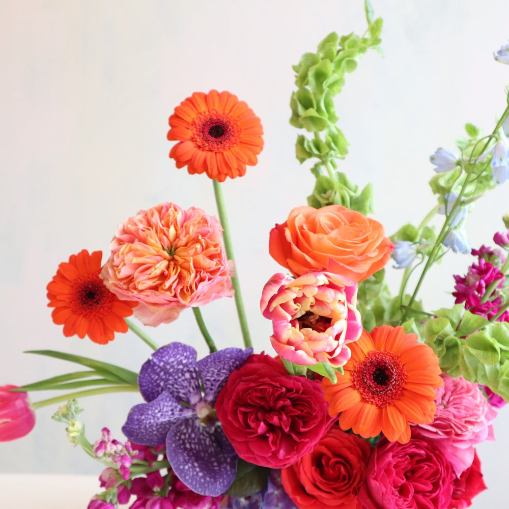 Bright and colorful floral arranegement, with orange gerbera daisies, bells of ireland, fushia garden roses, blue delphinium, purple vanda orchids, burgundy stock in a purple low compote.