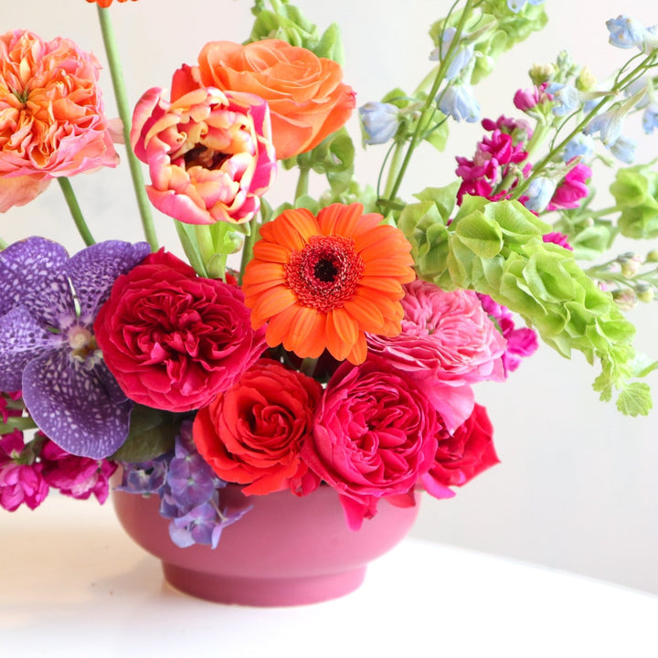 Bright and colorful floral arranegement, with orange gerbera daisies, bells of ireland, fushia garden roses, blue delphinium, purple vanda orchids, burgundy stock in a purple low compote.