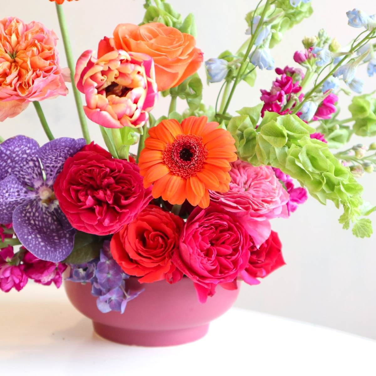 Bright and colorful floral arranegement, with orange gerbera daisies, bells of ireland, fushia garden roses, blue delphinium, purple vanda orchids, burgundy stock in a purple low compote.