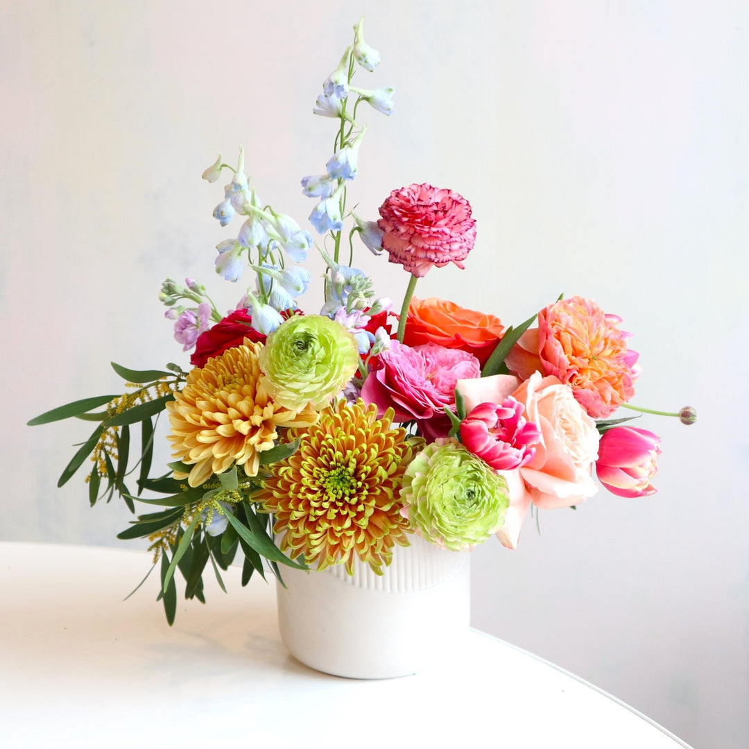 Flower arrangement with bronze mums, green ranunculus, pink garden roses, pink tulips, blue delphinium, greenery in a cream vase.