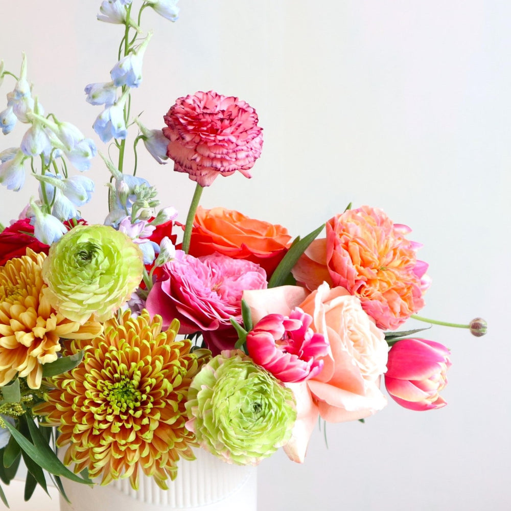 Flower arrangement with bronze mums, green ranunculus, pink garden roses, pink tulips, blue delphinium, greenery in a cream vase.
