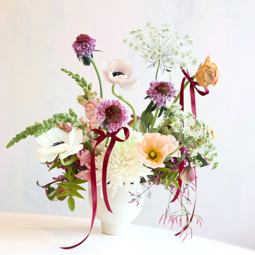 Graceful Garden | A fresh floral arrangement with purple, pink, white, and green. Accent ribbon is tied around select blooms. Made in a white vase.