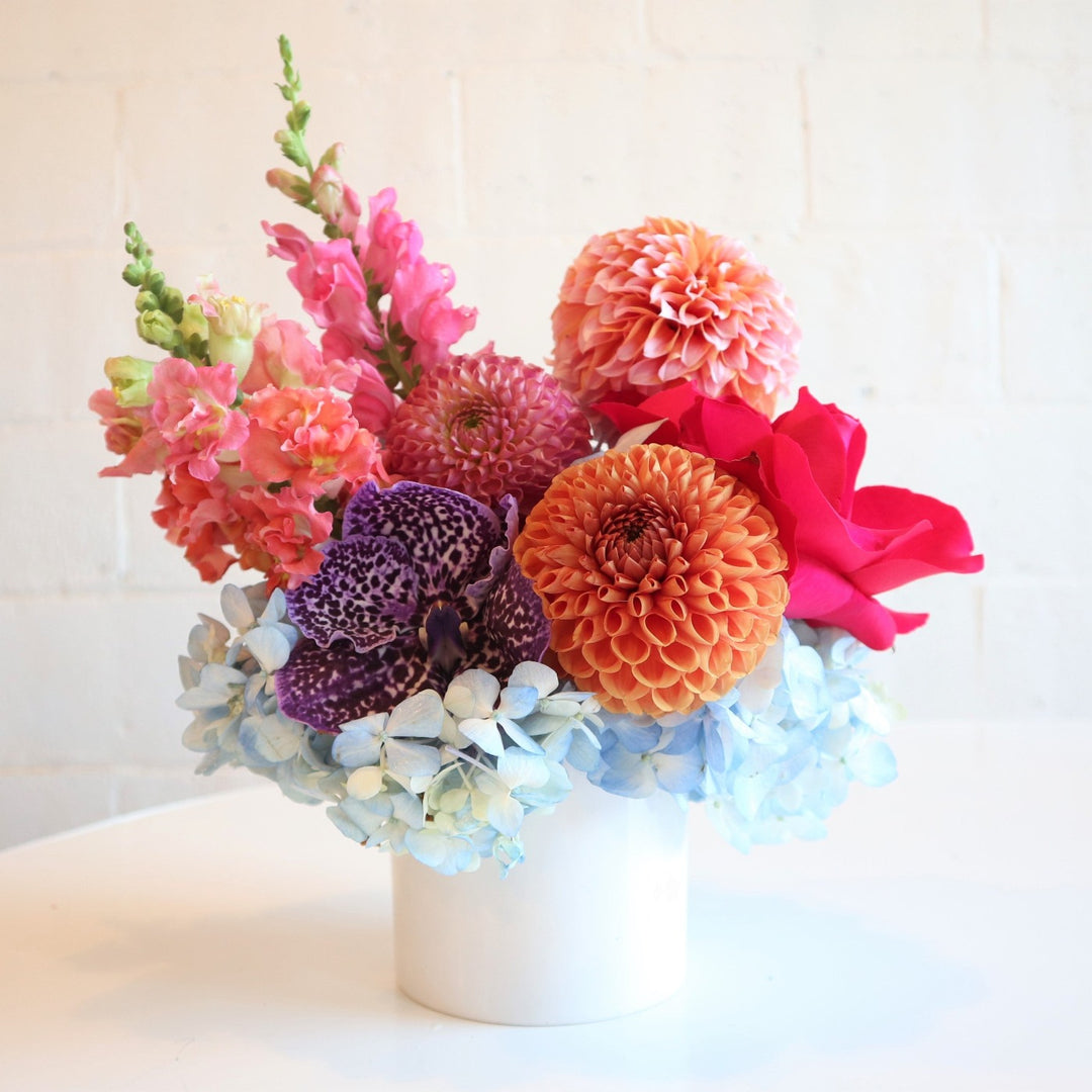 Cheerful | Vased Arrangement | A colorful floral arrangement with snapdragons, dahlias, hydrangea, and an orchid. In a white vase, photo taken against a white background. Flowers are pink, blue, purple, and orange.