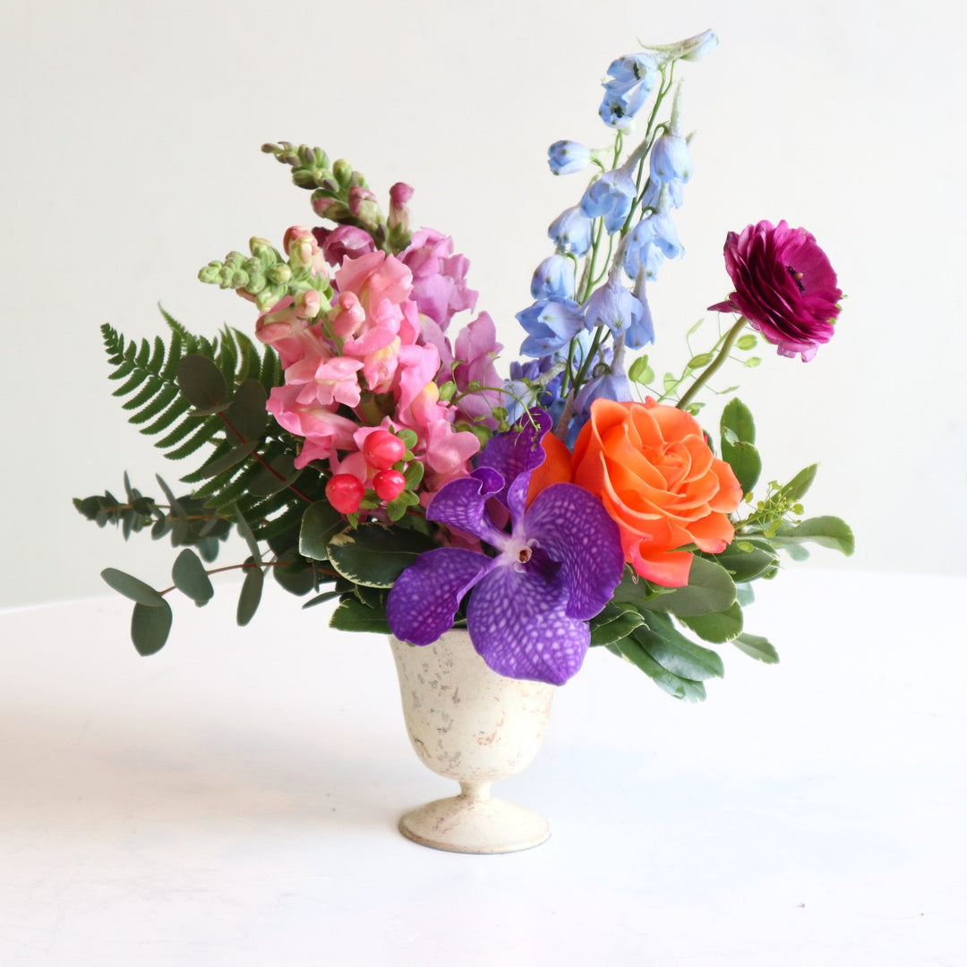 burst of color with purple snaps, blue delphinium, purple vanda orchids, and an orange rose in a cream vase.