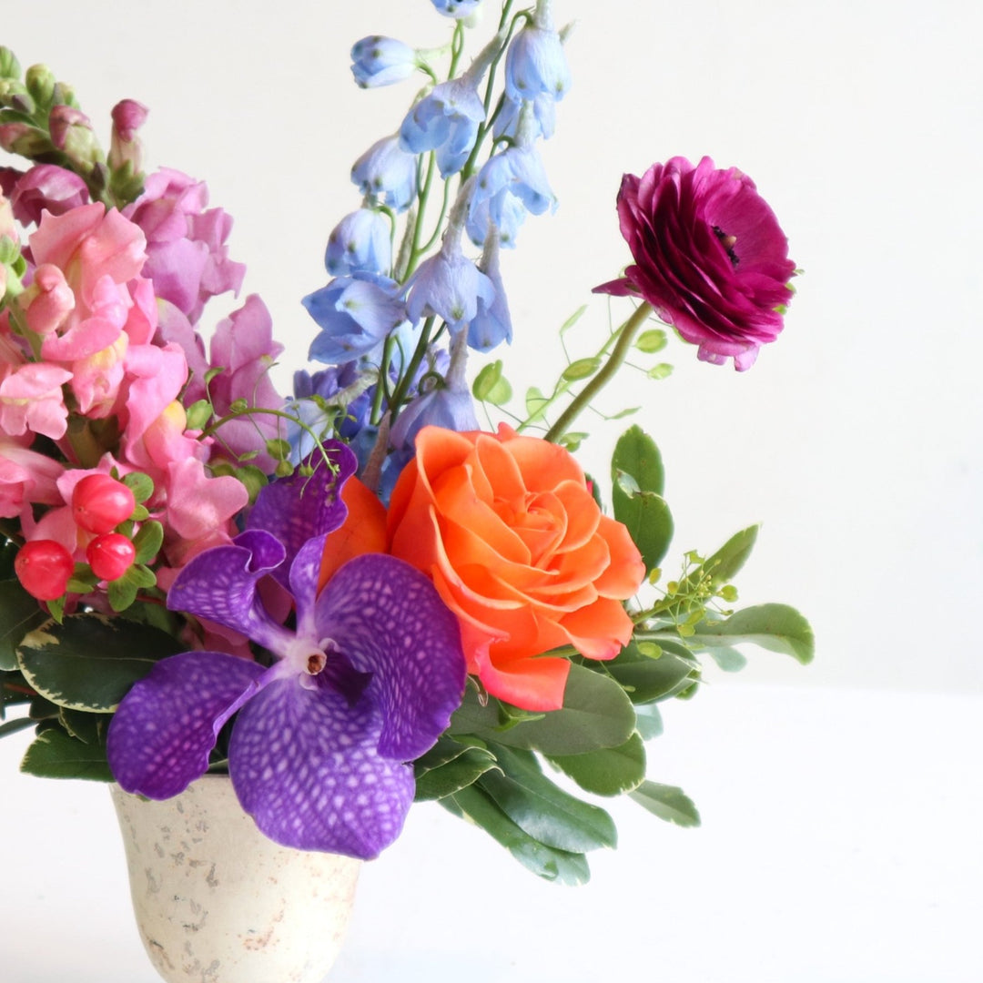 Close up of burst of color with purple snaps, blue delphinium, purple vanda orchids, and an orange rose in a cream vase.