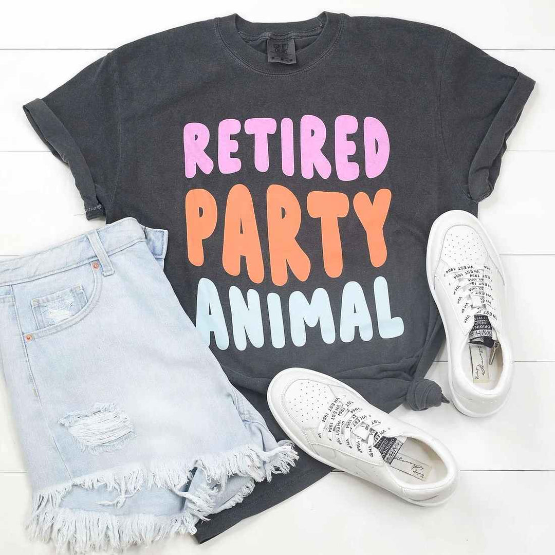 Mugsby | Retired Party Animal Shirt | A dark gray shirt with text in purple, orange, and blue that reads "Retired Party Animal". Photo shows the shirt, jean shorts, and white shoes.