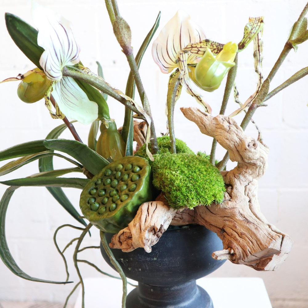 Faux flower arrangement with grapewood, lotus pods, lady slipper orchids, moss in a black vessel.