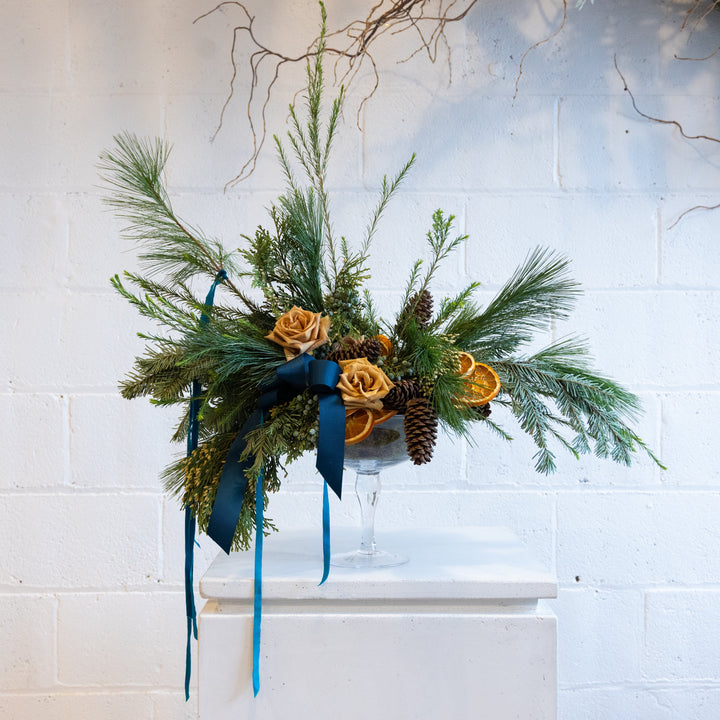 Decorative floral arrangement with greenery, orange flowers, and blue ribbons on a white surface against a light blue brick wall.