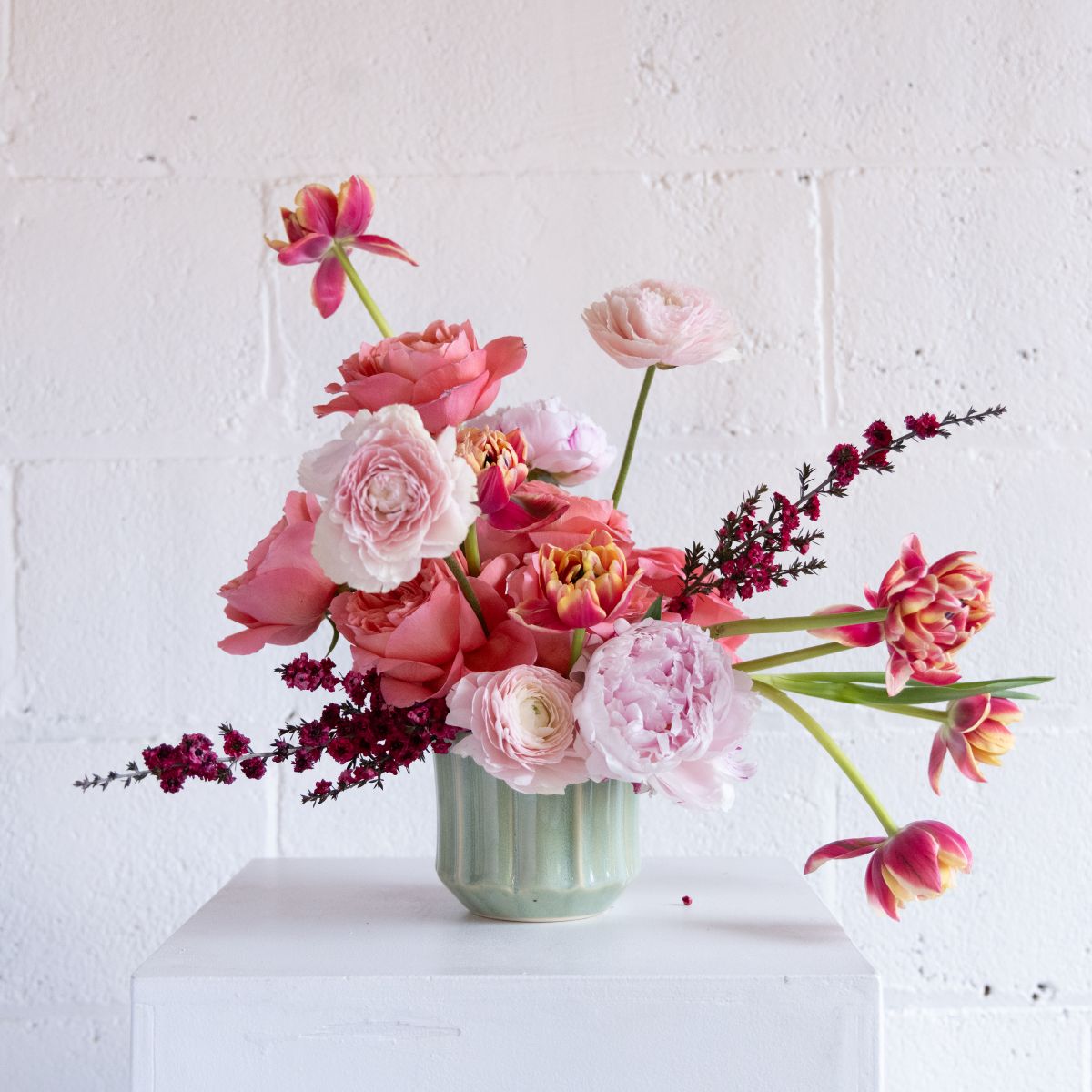 Bouquet of pink and white flowers in a green vase on a white surface with a white brick wall background.