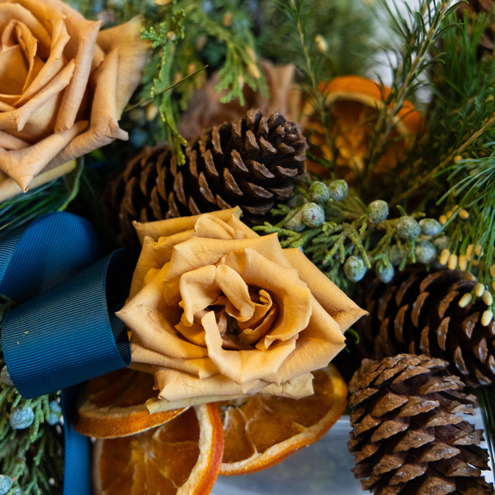 Decorative arrangement with beige roses, pine cones, and greenery on a reflective surface.
