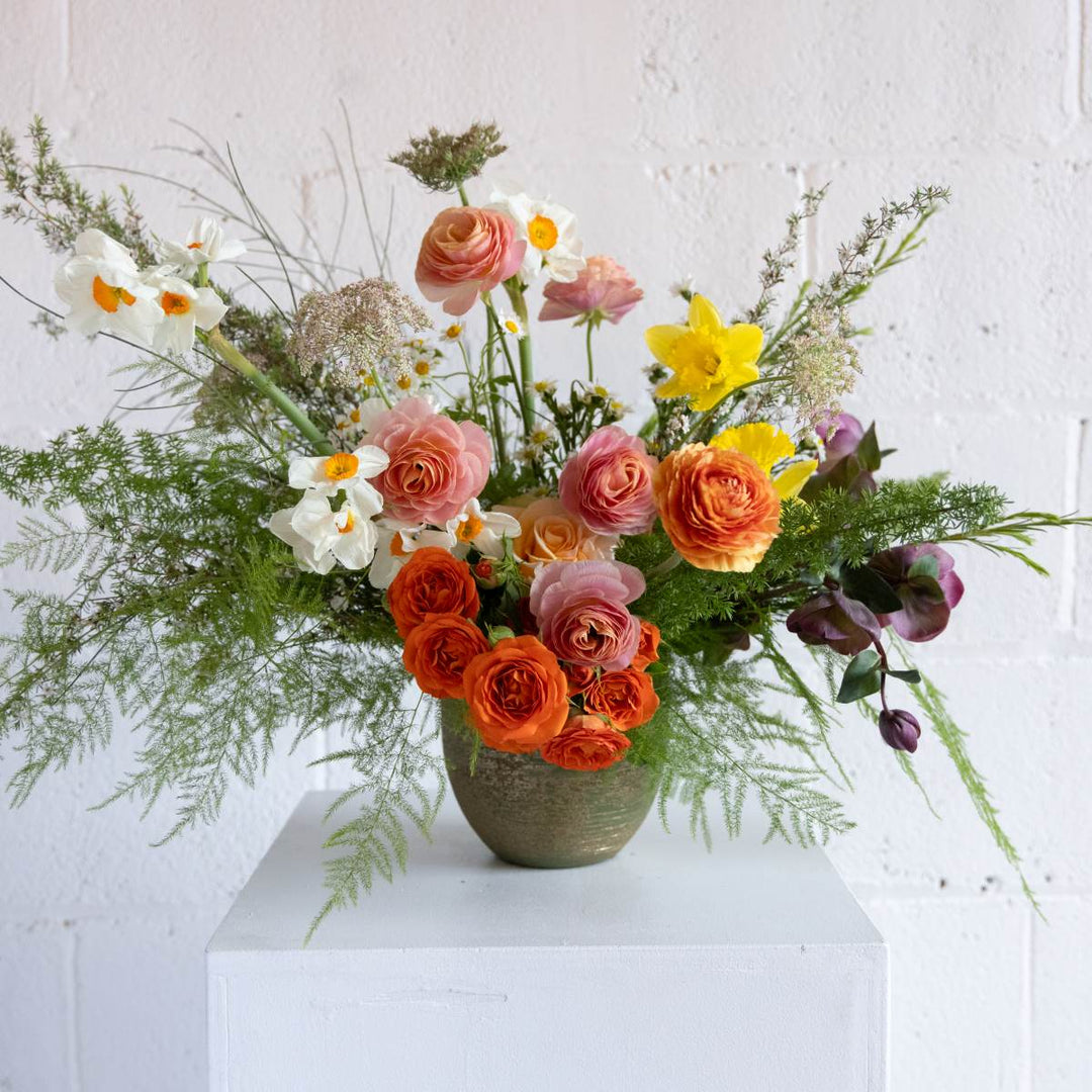 Colorful flower arrangement in a vase on a white surface with a light gray background, with roses, daffodils, greenery and more.