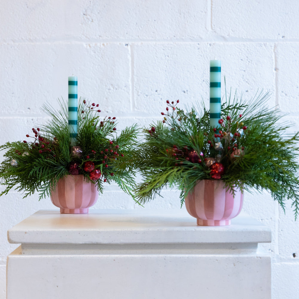 Two pink striped vases with greenery and red berries on a white surface against a white brick wall.