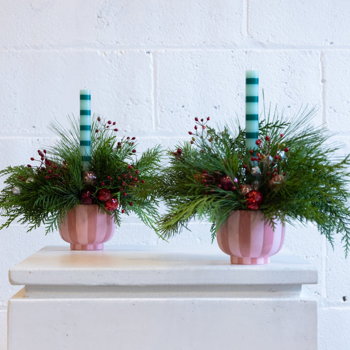 Two pink striped vases with greenery and red berries on a white surface against a white brick wall.