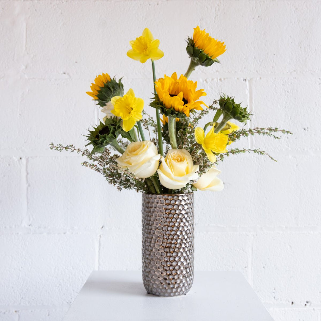 Floral arrangement with sunflowers and roses in a textured silver vase against a white brick wall.