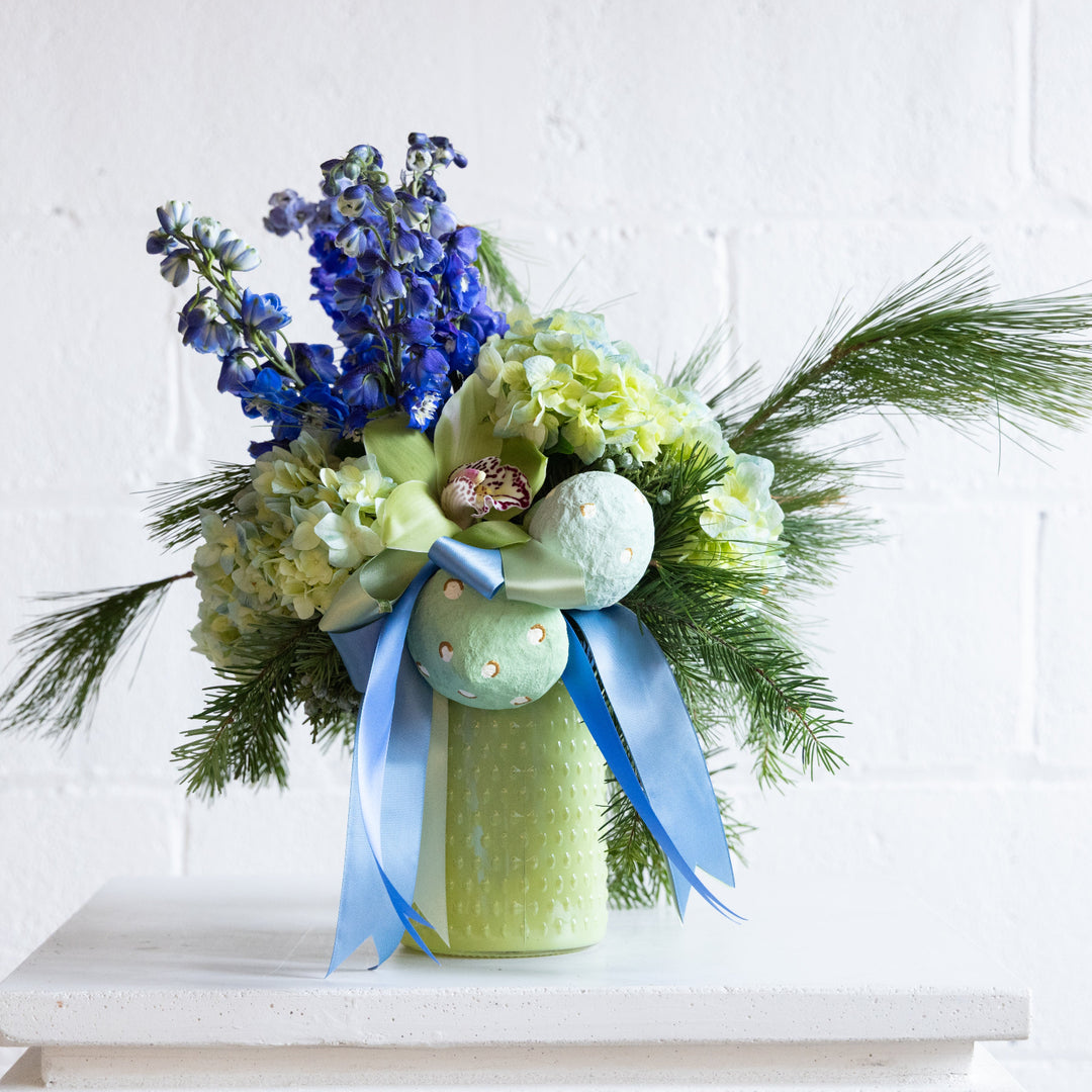 Decorative flower arrangement with blue and green ribbons in a green vase on a white surface.