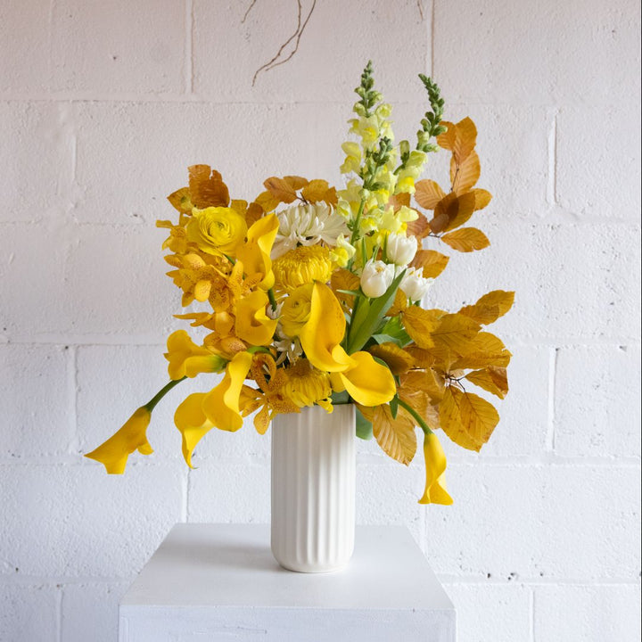 Bouquet of yellow and white flowers in a white vase against a white brick wall.