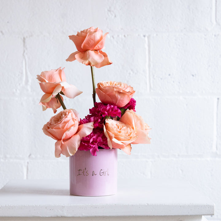 Pink flowers in a vase with 'It's a Girl' text against a white brick wall.