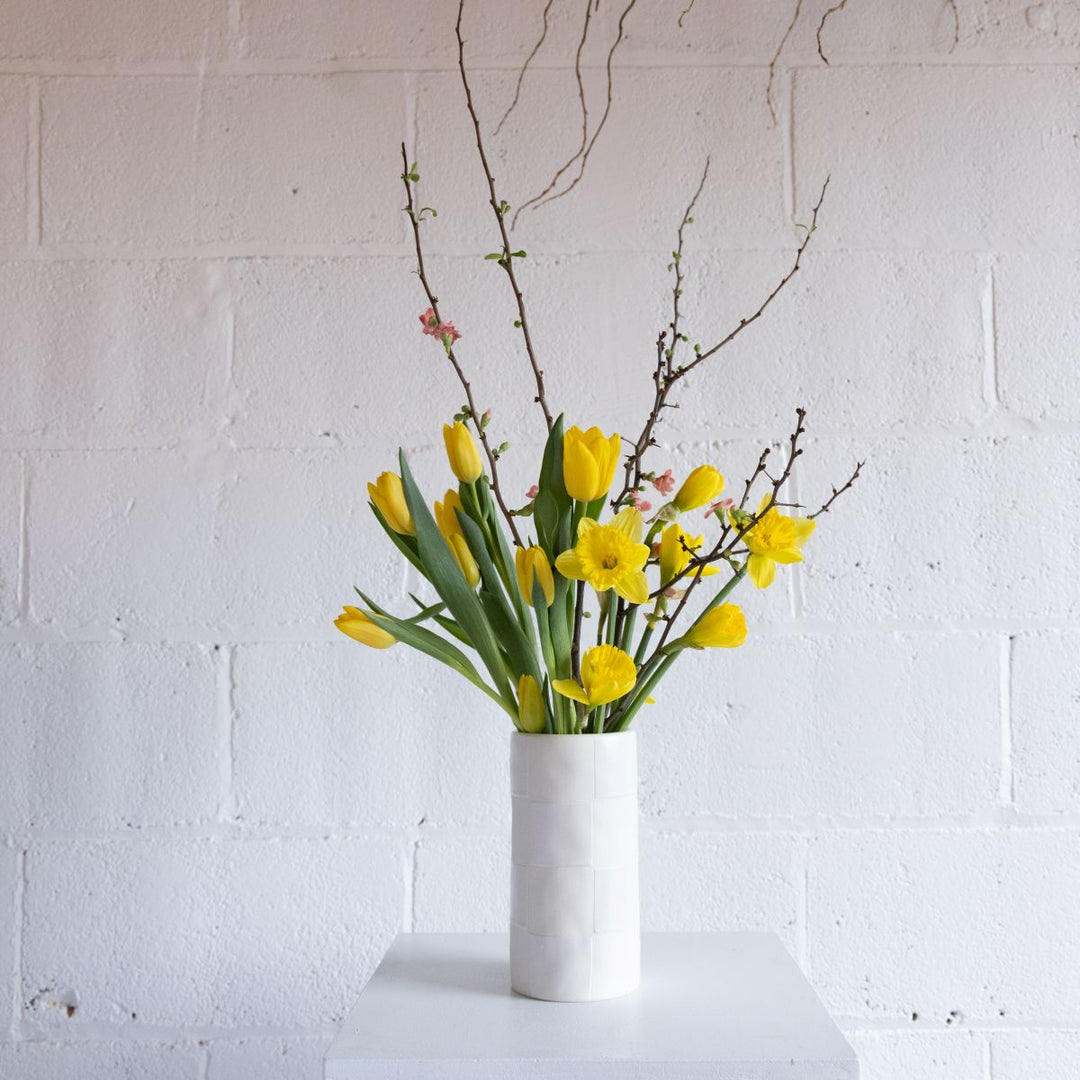 White vase with yellow flowers and green leaves against a white brick wall