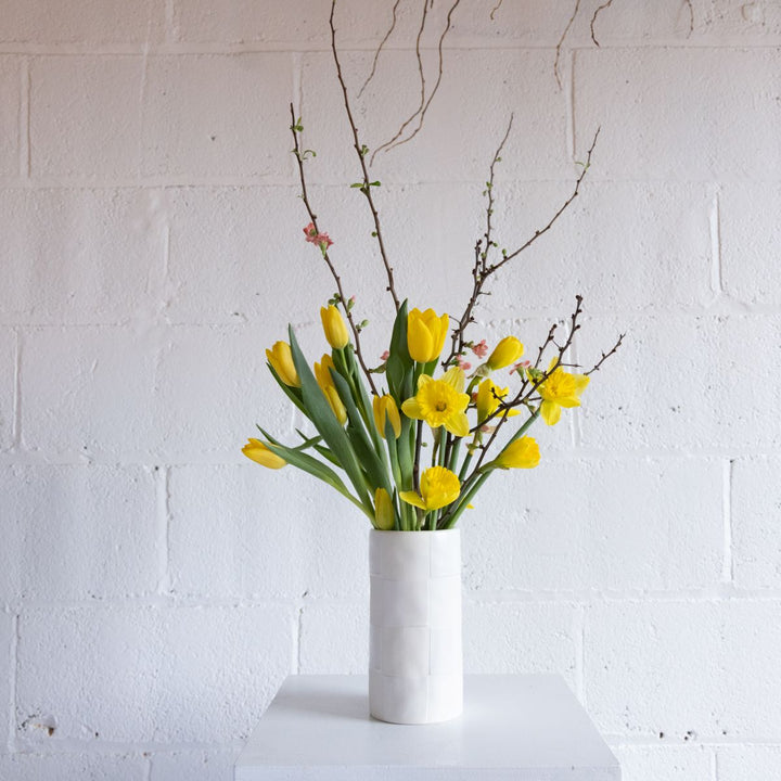 White vase with yellow flowers and green leaves against a white brick wall