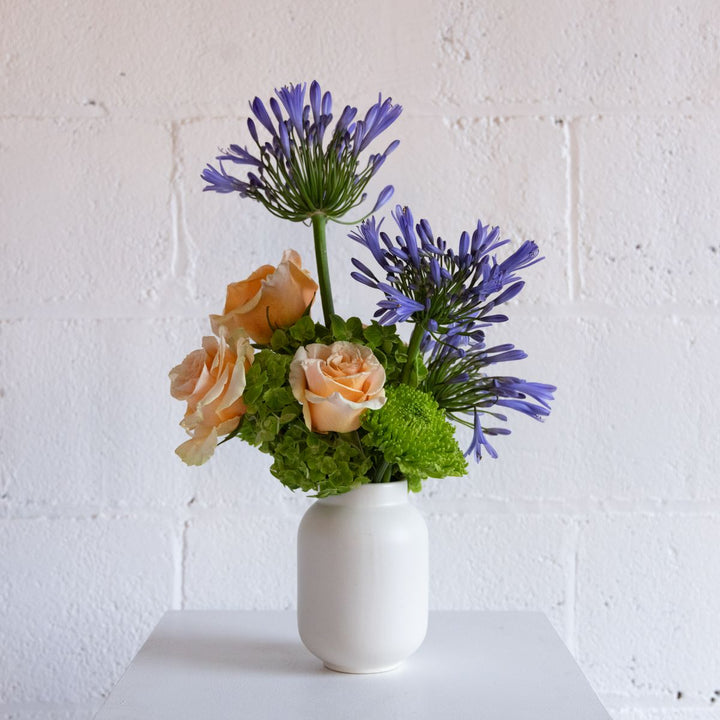 Floral arrangement with purple flowers and peach roses in a white vase against a white brick wall.