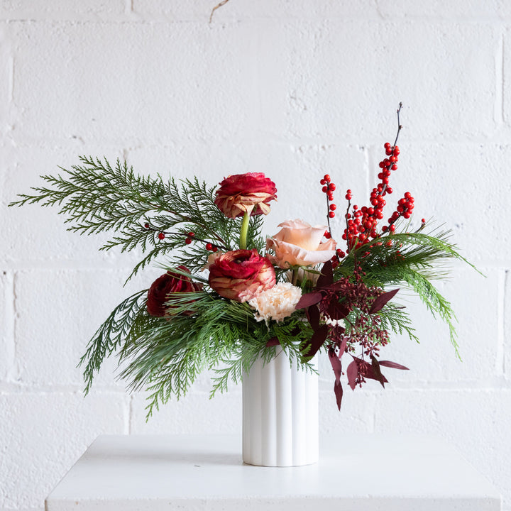 Floral arrangement with red and green elements in a white vase against a white brick wall.