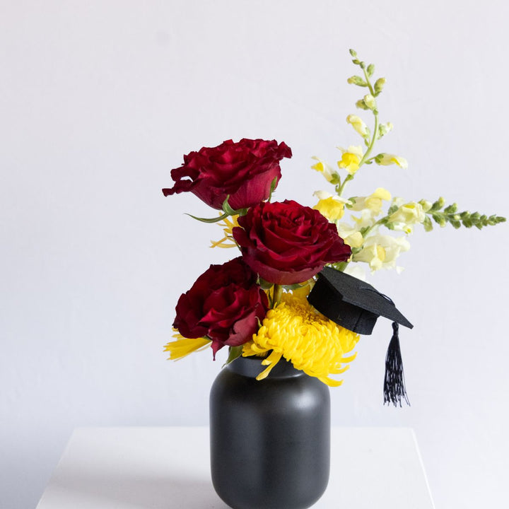 Black vase with red roses, yellow flowers, and a small graduation cap on a light gray background