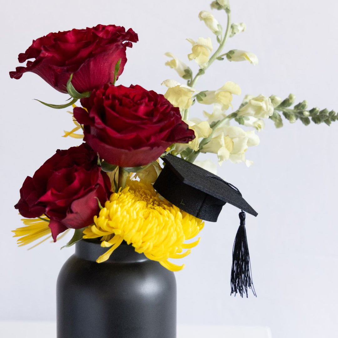 Vase with red roses and yellow flowers, featuring a small graduation cap with tassel.