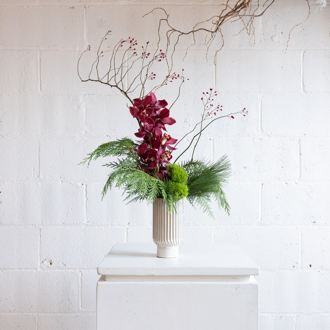Floral arrangement with red flowers and greenery in a white vase against a white brick wall.