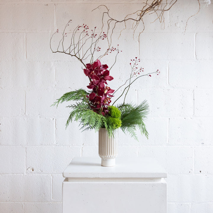 Floral arrangement with red flowers and greenery in a white vase against a white brick wall.