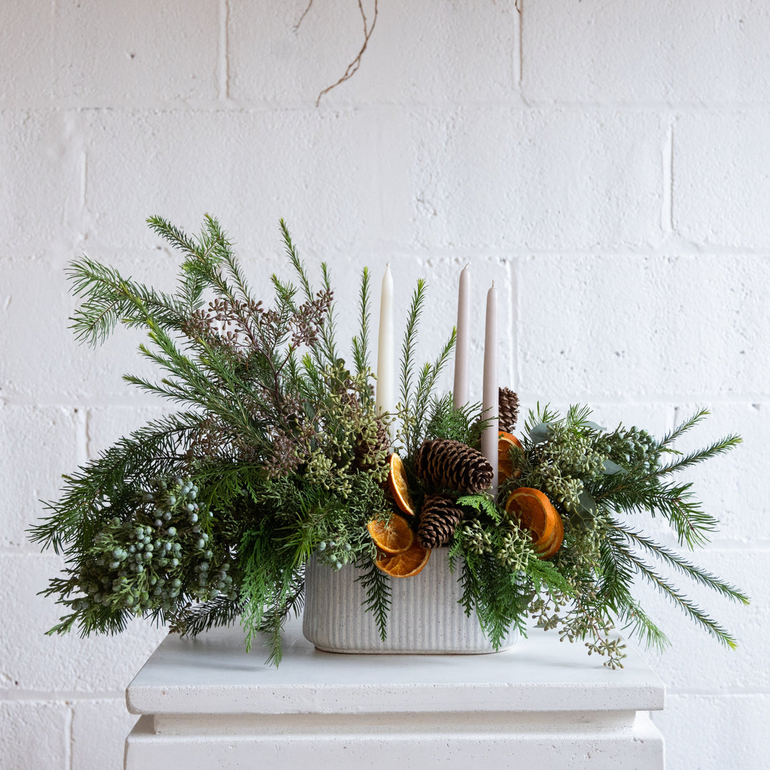 Decorative arrangement with greenery, pinecones, and candles on a white surface against a white brick wall.