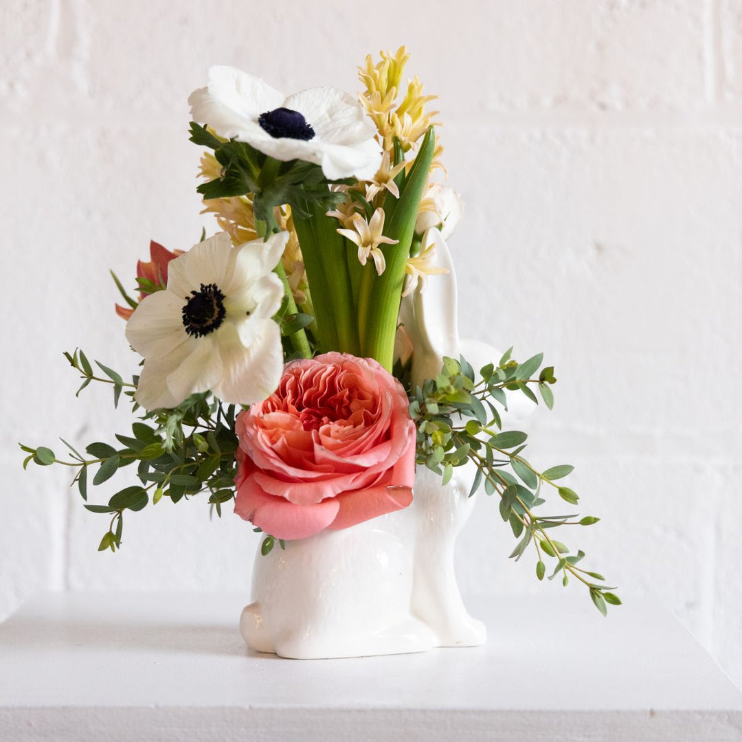 Floral arrangement with pink rose, white anemone, and green leaves in a white bunny vase on a light background