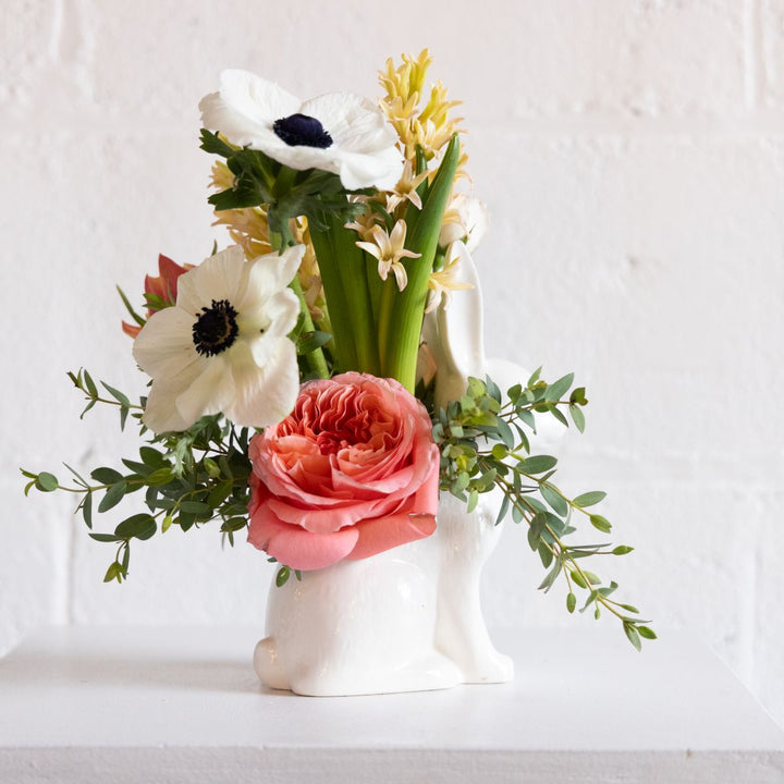 Floral arrangement with pink rose, white anemone, and green leaves in a white bunny vase on a light background