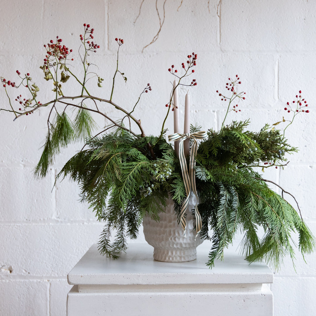 Decorative arrangement with greenery, candles, and ribbons on a white surface against a white brick wall.