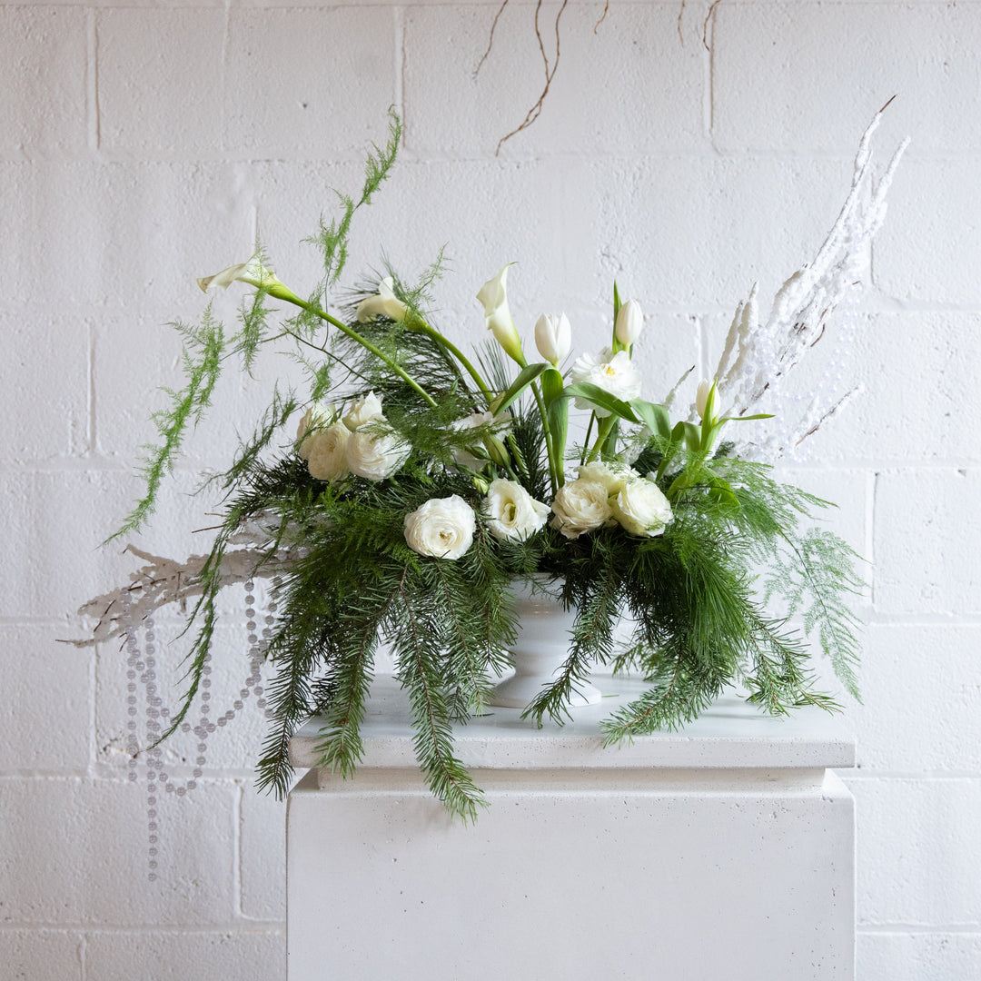 Floral arrangement with white flowers and greenery on a white surface against a light gray brick wall.