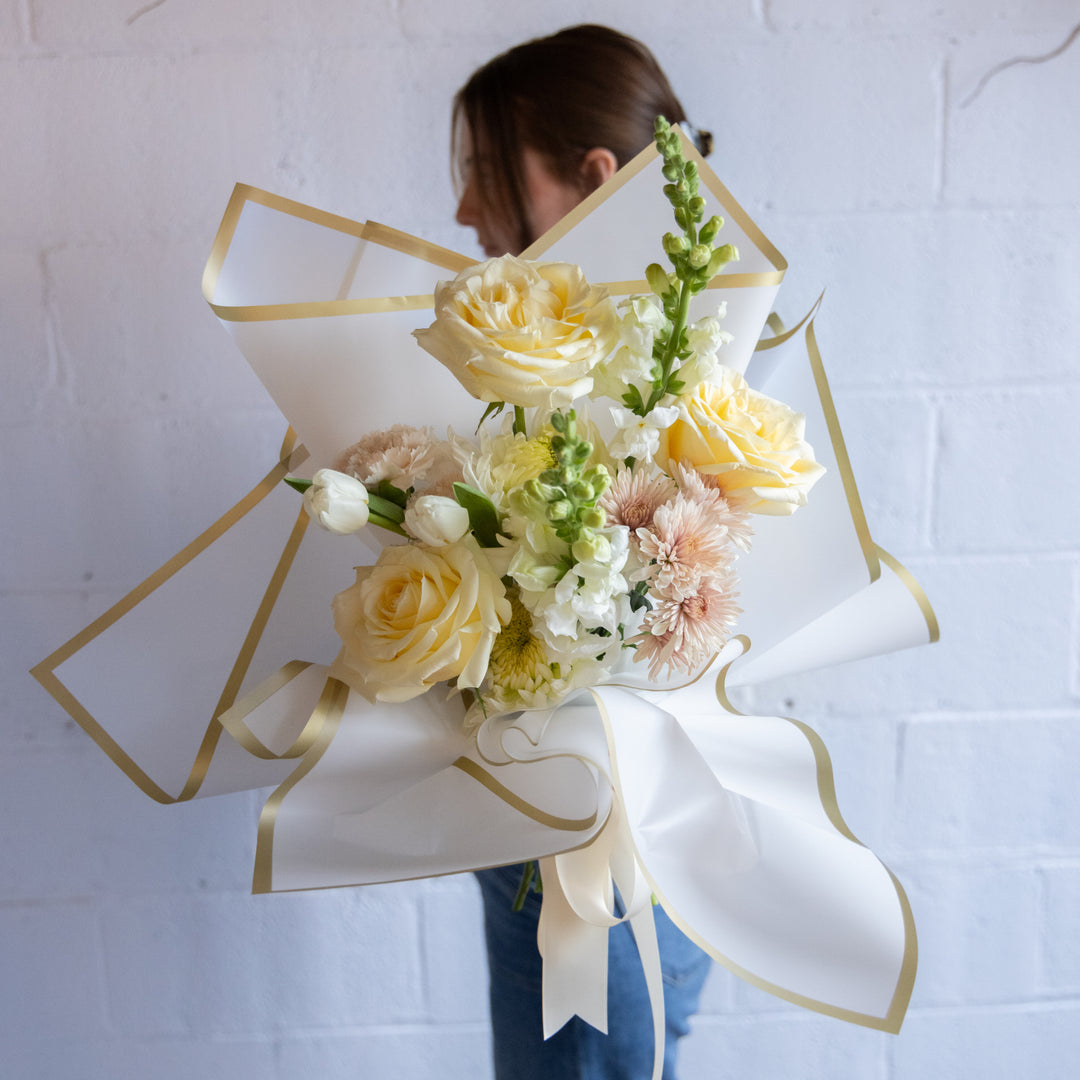 Bouquet of flowers wrapped in white paper with gold ribbons held by a person against a white background