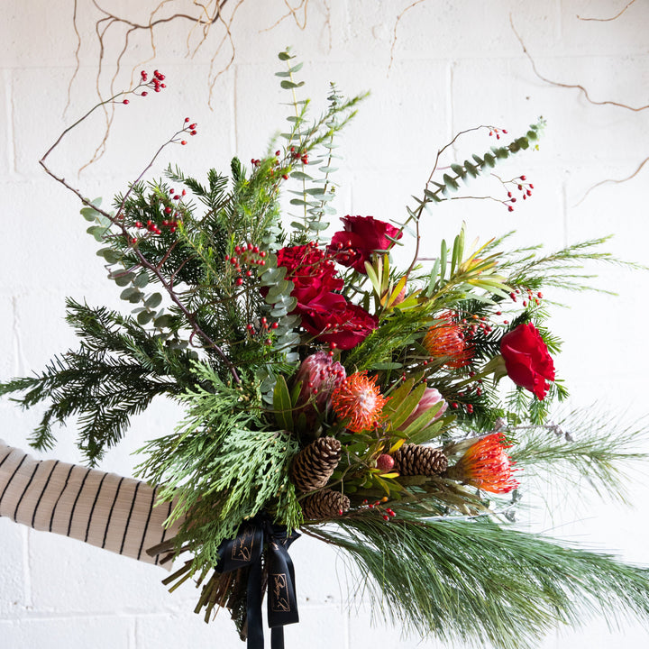 Bouquet of flowers with red roses, greenery, and pinecones held by a person against a light background.