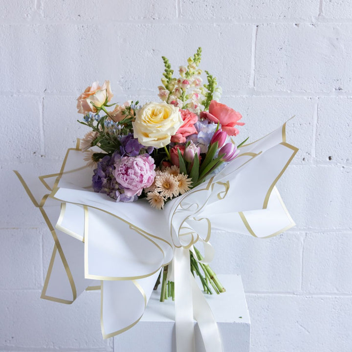 Bouquet of flowers wrapped in white paper with gold accents against a light gray brick wall.
